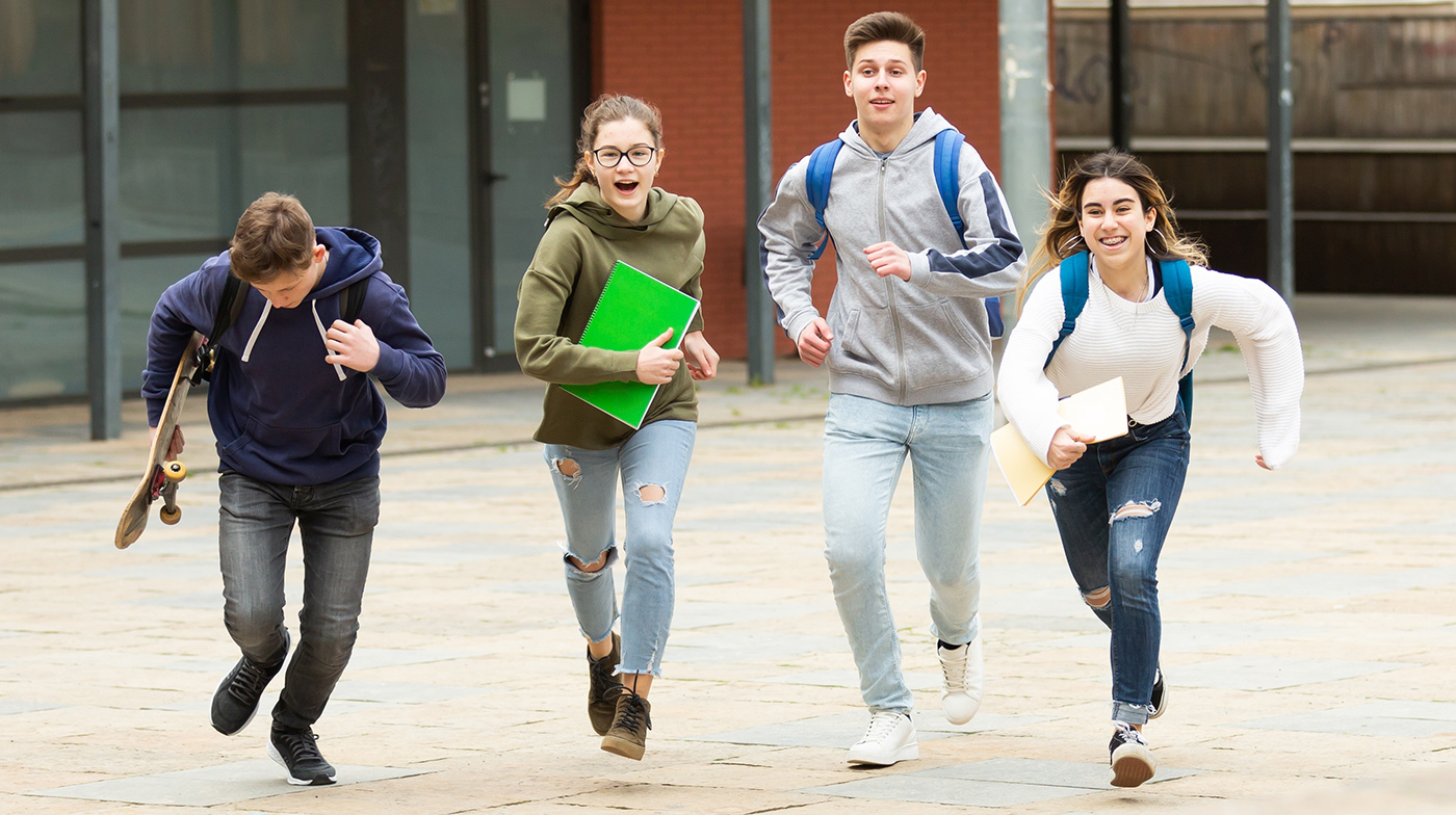 Four teenagers, two boys and two girls, are smiling and running outdoors. One boy holds a skateboard, and the others carry notebooks or folders. They appear happy and energetic, dressed casually with backpacks.