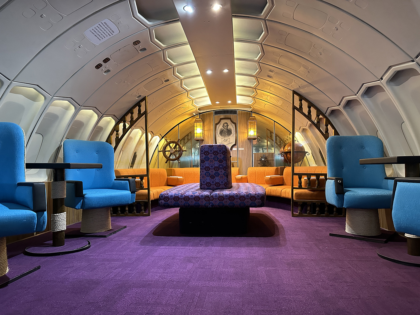 A stylish lounge inside an airplane fuselage, featuring blue chairs, orange sofas, a purple carpet, and nautical decor elements. Warm lighting creates a cozy, retro-modern atmosphere.