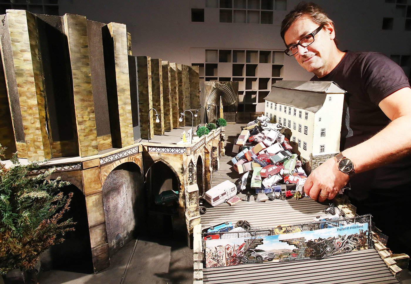 A man arranges a detailed miniature model of a cityscape featuring a bridge, trees, buildings, streetlights, and piles of tiny cars, with urban graffiti painted along a wall in the foreground.