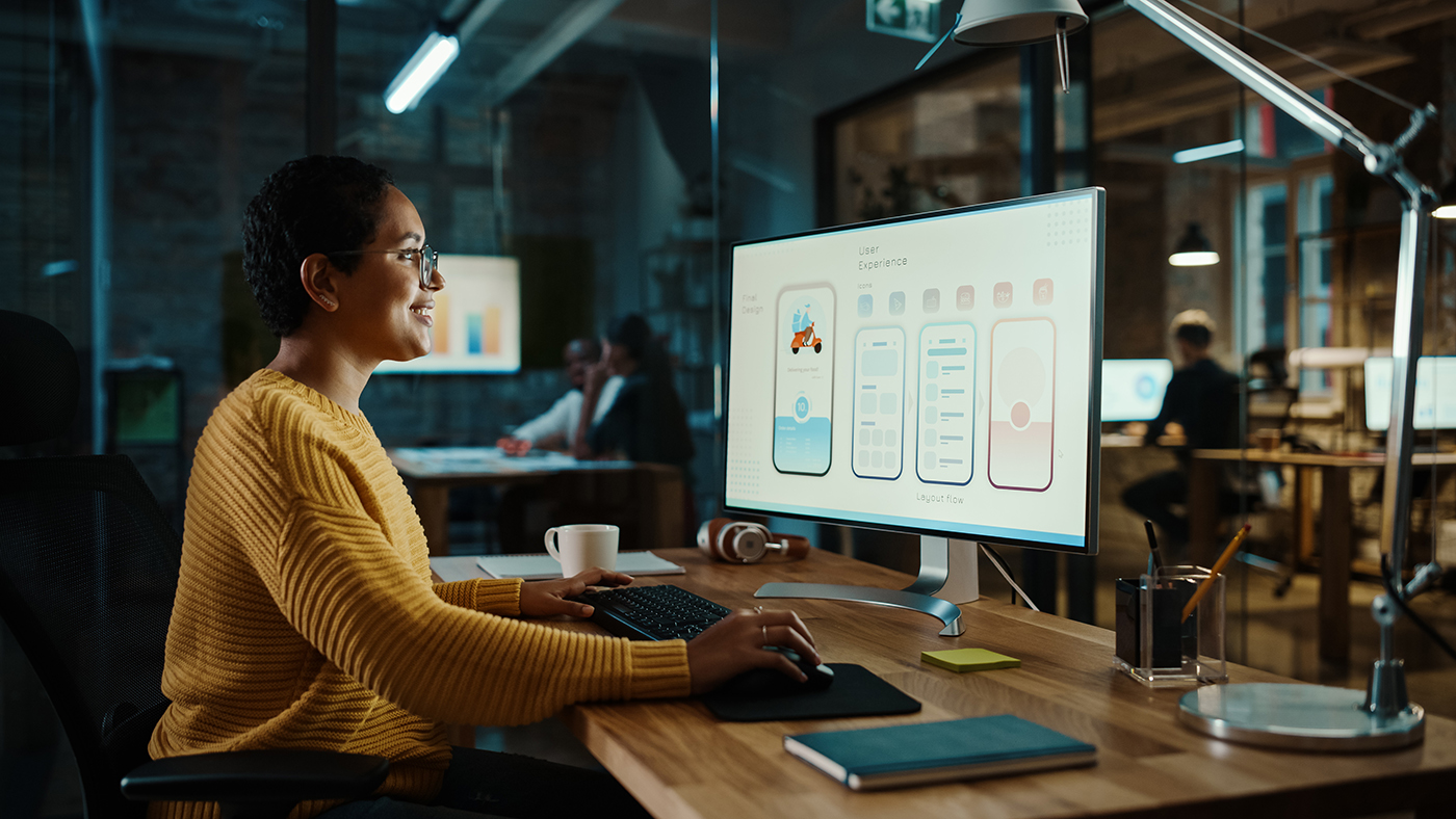 A person in a yellow sweater sits at a desk in a modern office, working on a computer displaying user experience design screens. Office supplies and a lamp are on the desk, with colleagues visible in the background.