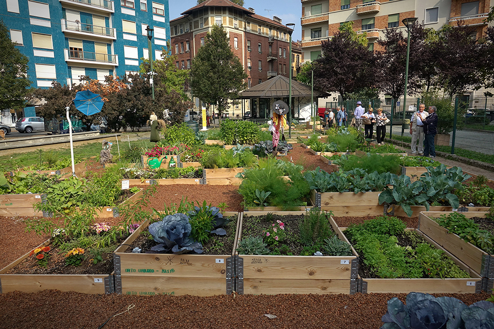 An urban community garden with raised wooden beds filled with vegetables and plants. People are walking and gathering in the background, surrounded by apartment buildings and trees on a sunny day.