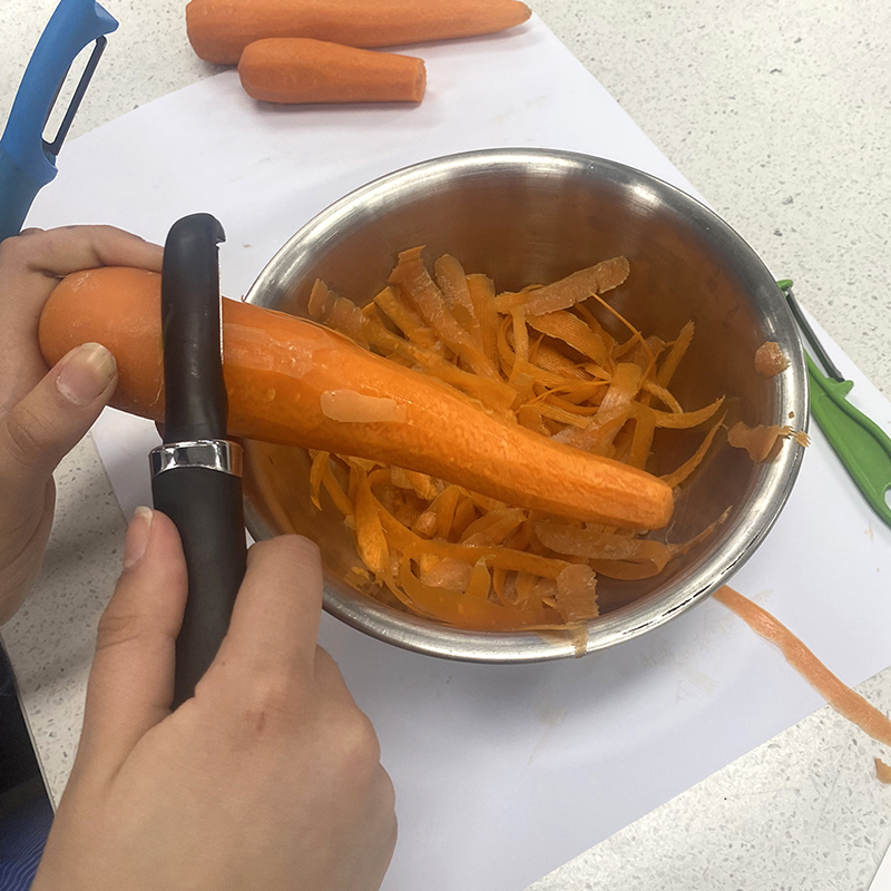 A person uses a vegetable peeler to peel a carrot over a metal bowl filled with carrot peels. Other unpeeled carrots and a green peeler are on the counter nearby.
