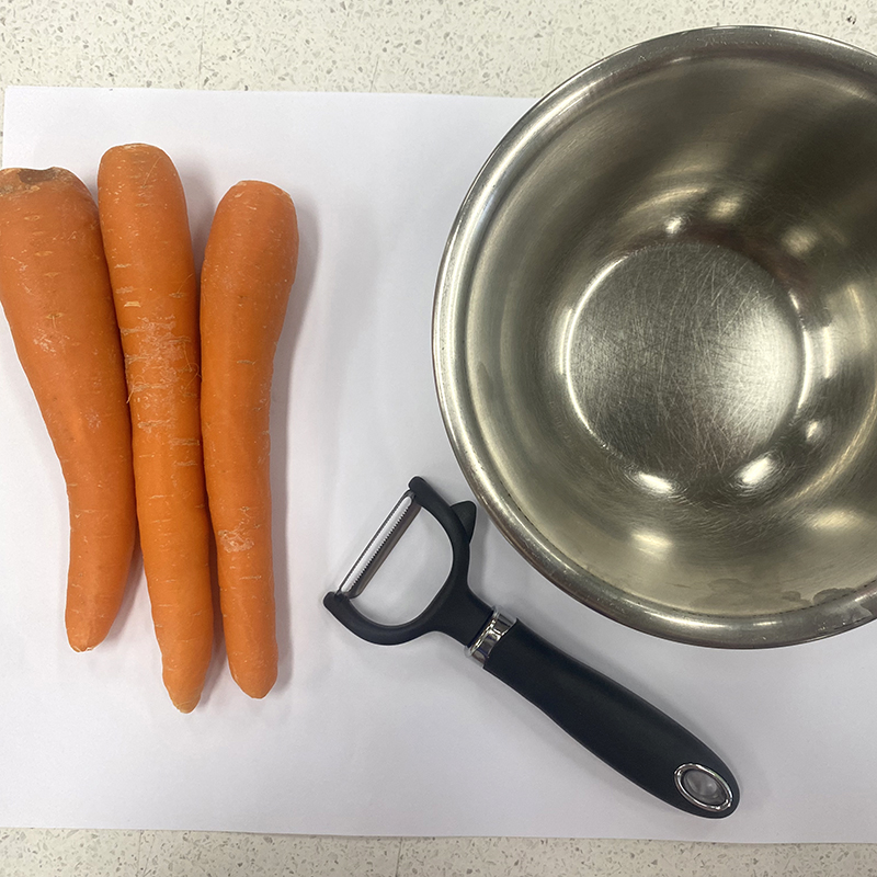 Three whole carrots, a vegetable peeler, and a metal mixing bowl are placed on a white sheet of paper on a light-colored countertop.