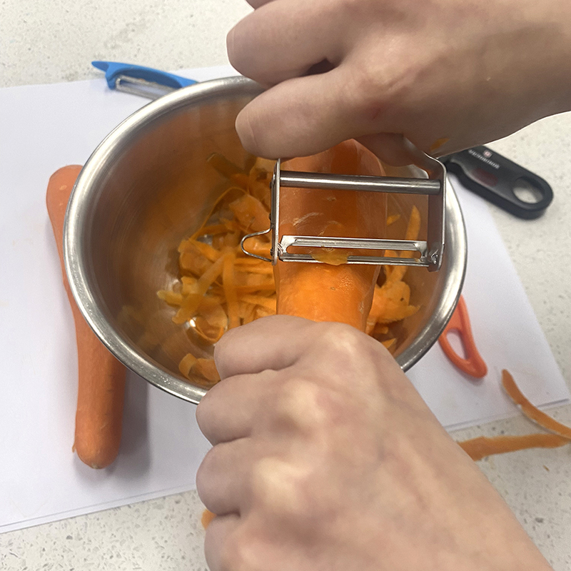 A person peeling a carrot over a metal bowl filled with carrot peels; another carrot and a peeler are on the countertop nearby.