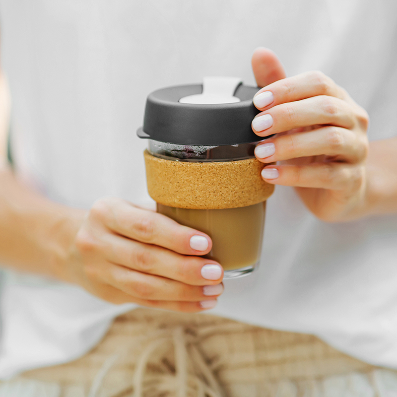 A person holding a reusable glass coffee cup with a cork band and a black lid, wearing a white shirt and beige pants. Their nails are painted light pink.
