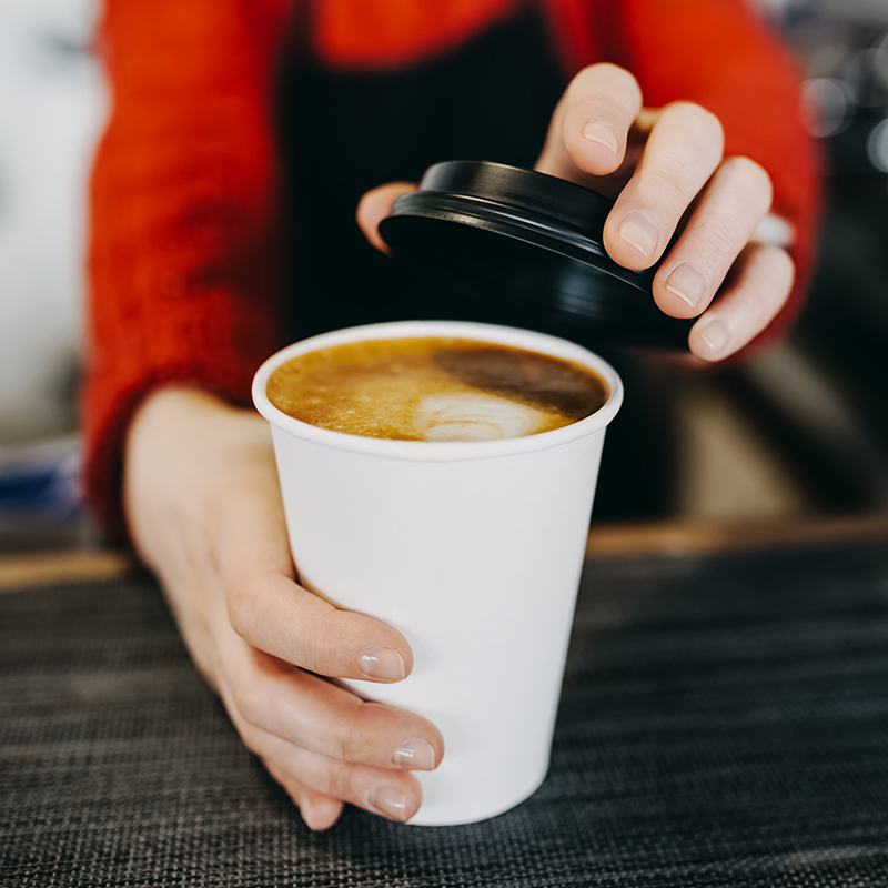 A person in a red sweater holds a white disposable coffee cup and is placing a black plastic lid on it. The cup is filled with coffee and some foam can be seen on top.