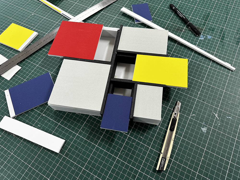 A geometric model with red, yellow, blue, white, and black rectangles sits on a green cutting mat. Surrounding it are a ruler, a hobby knife, a pen, and various strips of colored paper or card.