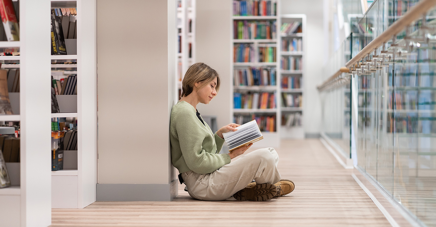 A person sits on a wooden floor, leaning against a white wall in a library, reading a book. Bookshelves filled with colorful books line the background. The setting is calm and well-lit.