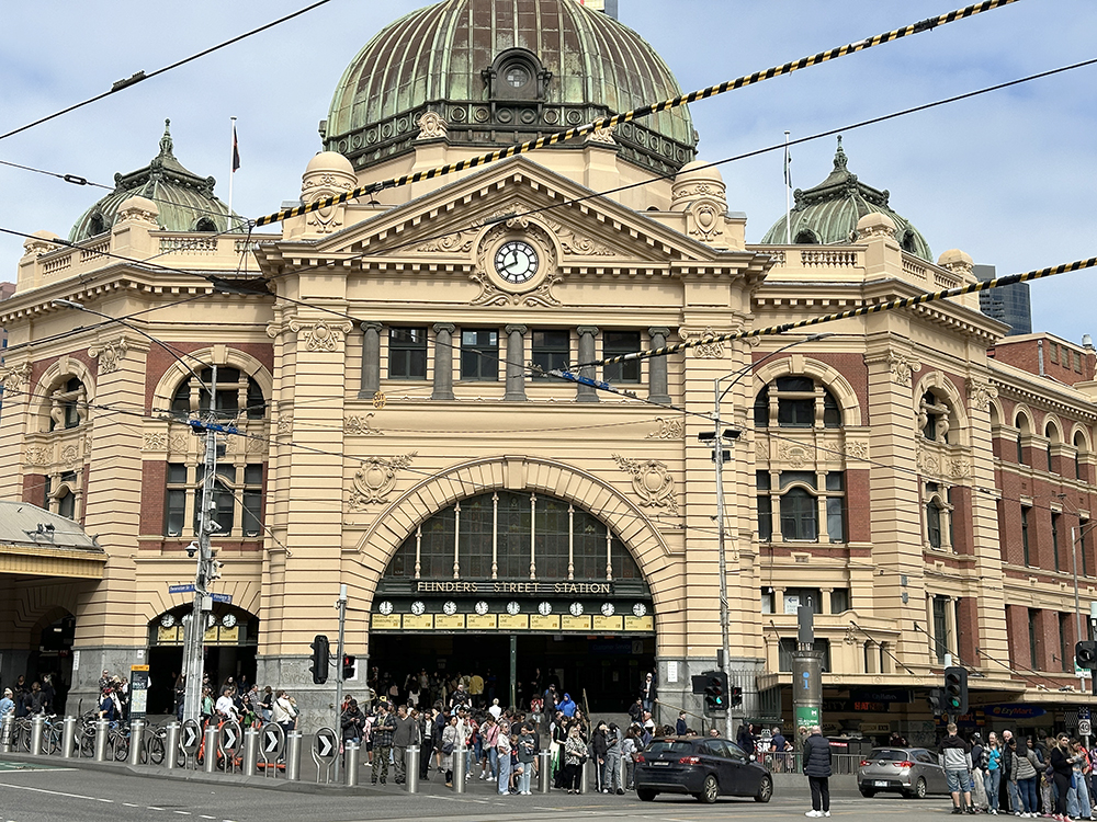 A busy street scene outside Flinders Street Station in Melbourne, with crowds of people, cars, trams, and the station’s iconic domed building under a partly cloudy sky.