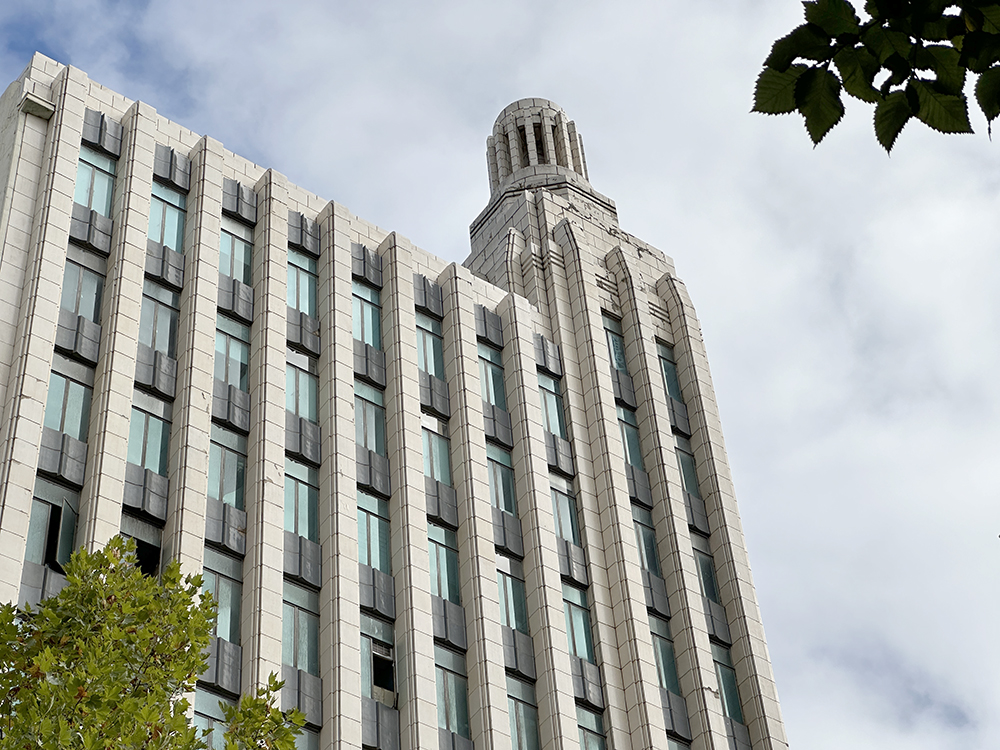 A tall, modern building with vertical windows and a distinctive rounded tower rises against a cloudy sky, with green tree branches in the foreground.