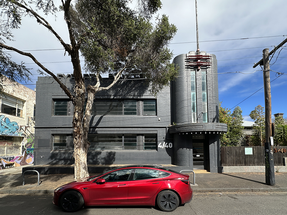 A red car is parked on the street in front of a modern gray brick building with horizontal windows and a curved entrance. A large tree stands between the car and the building. The sky is partly cloudy.