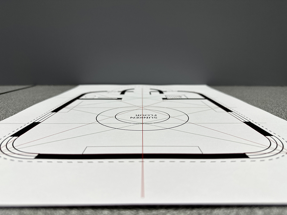 A close-up, low-angle view of a printed architectural or floor plan labeled SUNKEN FLOOR, with red grid lines and bold black outlines, placed on a gray surface against a plain gray background.