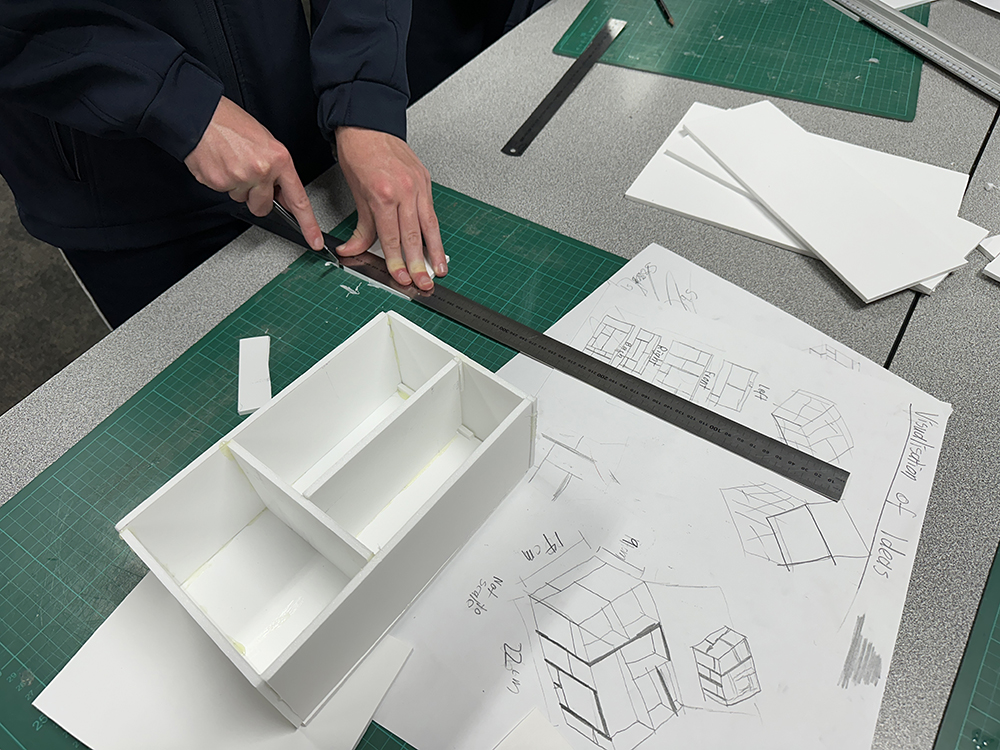 A person uses a metal ruler and utility knife to cut white foam board on a green cutting mat beside an architectural model and sketches on a table.