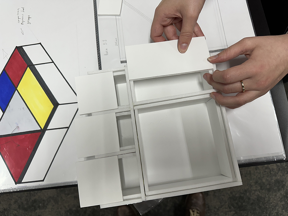 A person assembles a white architectural model with rectangular sections; a Mondrian-style drawing with red, blue, and yellow squares is visible on the table nearby.