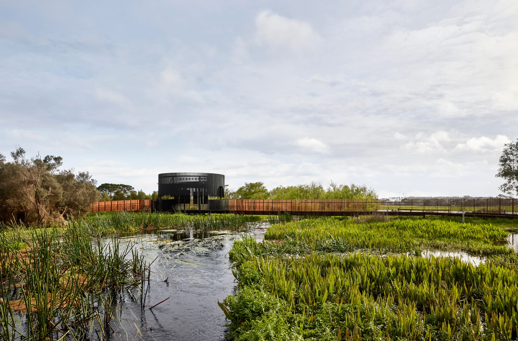 A modern, circular black building is set behind a wooden fence, surrounded by lush wetlands with tall grass and water plants under a cloudy sky. A wooden boardwalk leads through the natural landscape.