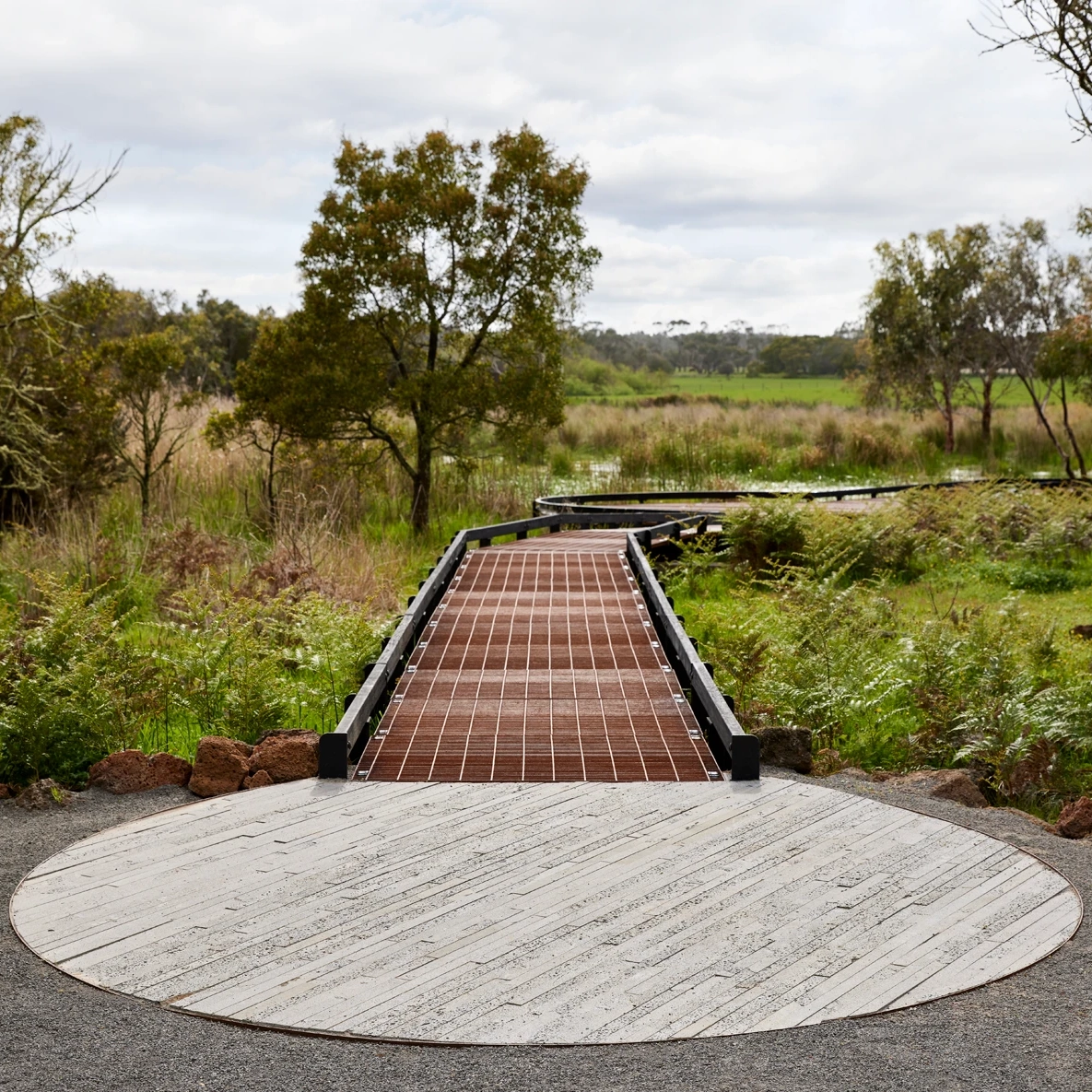 A wooden boardwalk with black railings curves through lush green grass and trees, starting from a round concrete platform in the foreground under a cloudy sky.