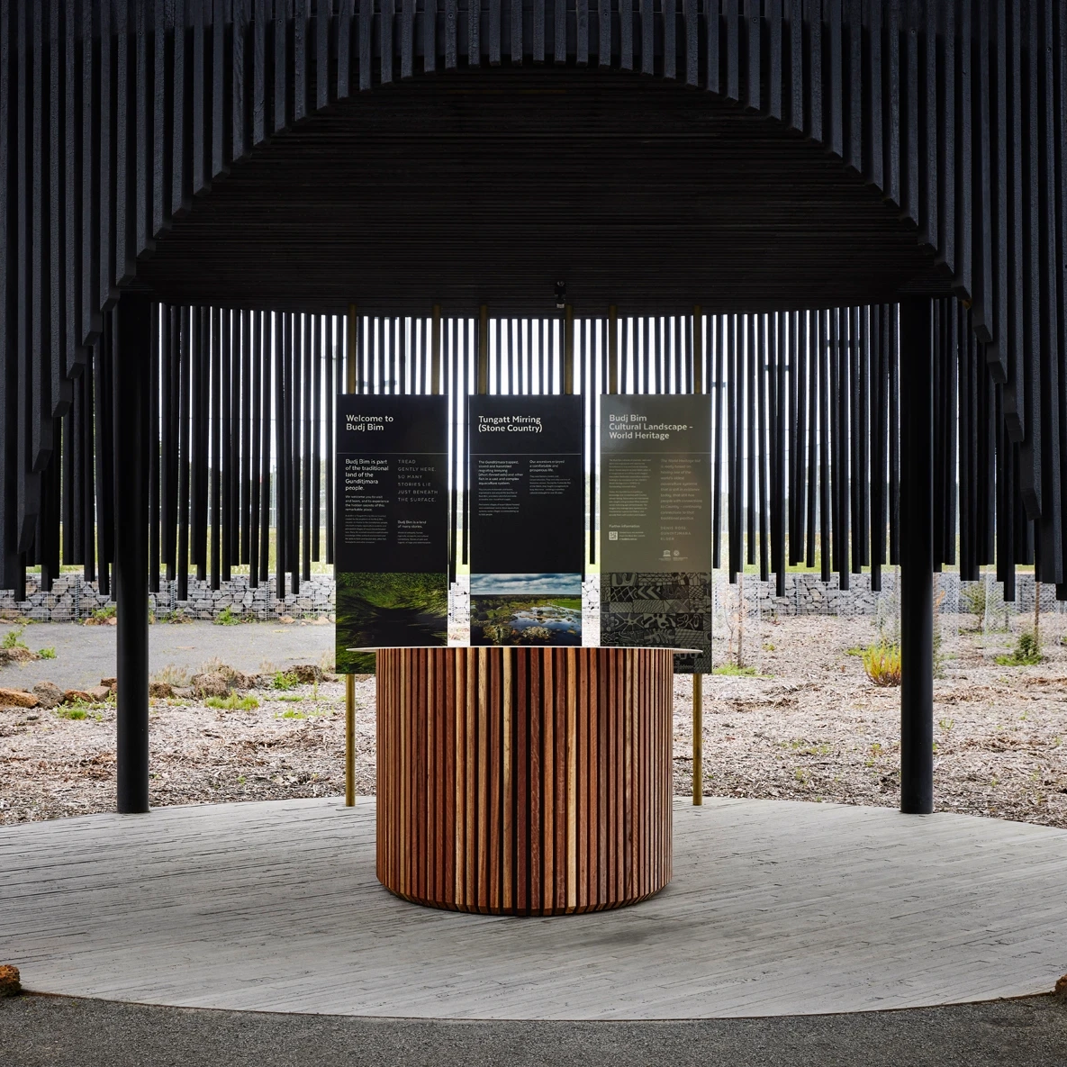 A modern outdoor information kiosk with vertical black slats, a round wooden table in front, and three informational panels displayed above, set against a gravel and shrub landscape.