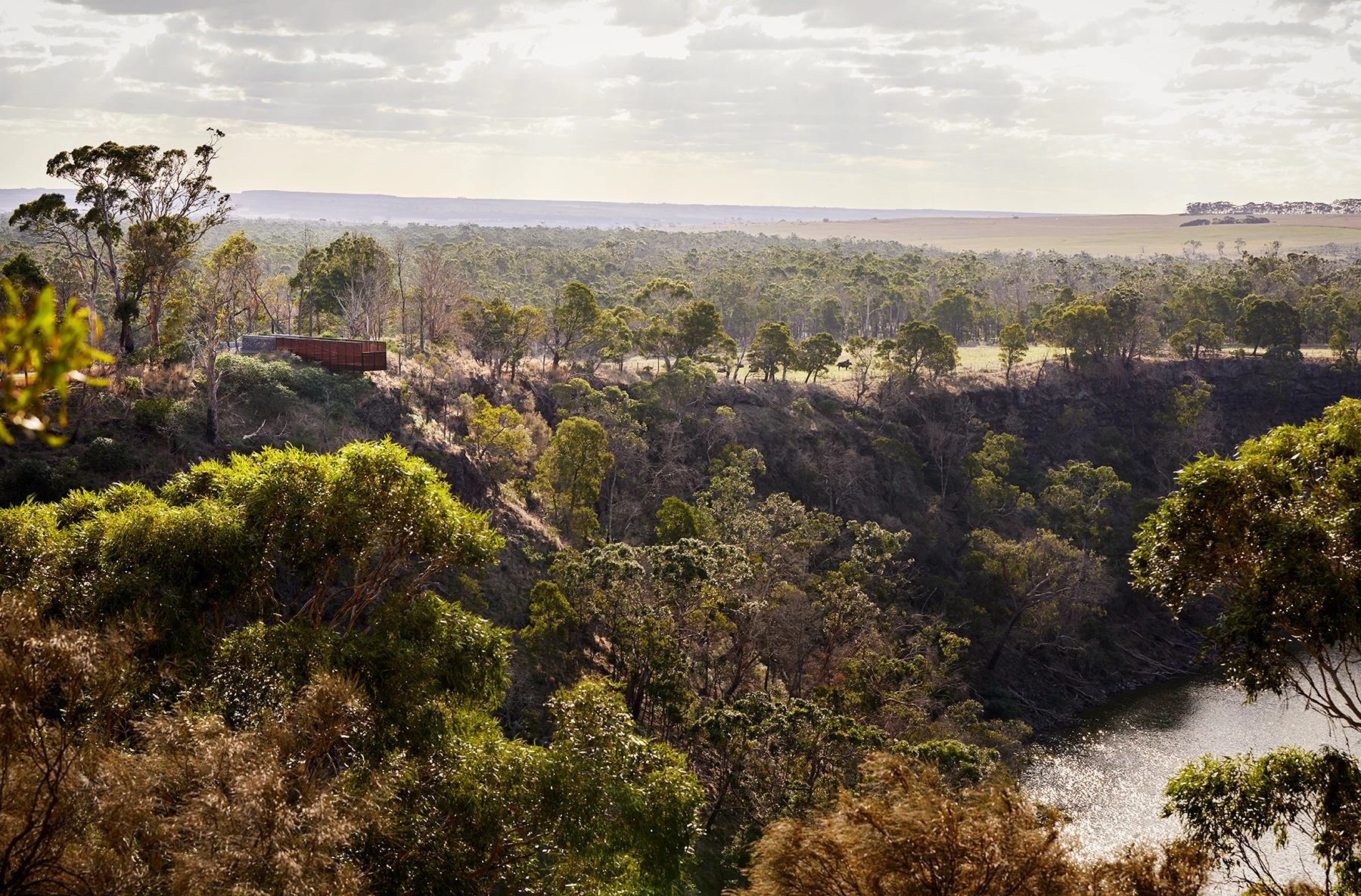 A scenic view of a lush green gorge with a river running at the bottom. Tall trees cover the landscape, and a small cabin is nestled among the trees. The sky is partly cloudy with sunlight streaming through.