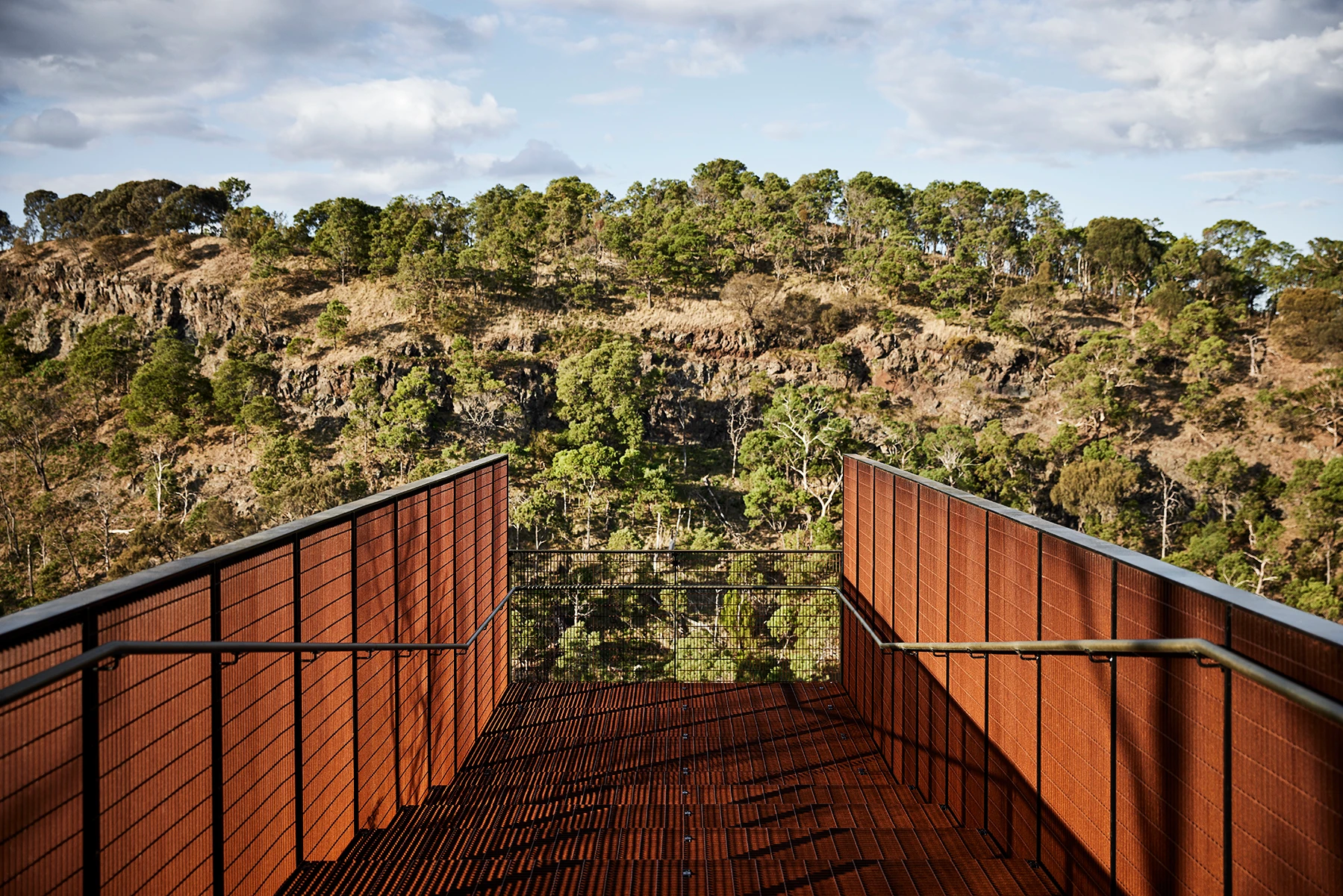 A rust-colored metal viewing platform with railings extends towards a rocky, tree-covered hillside under a partly cloudy sky. The platform is empty and offers a scenic, elevated view of the surrounding landscape.
