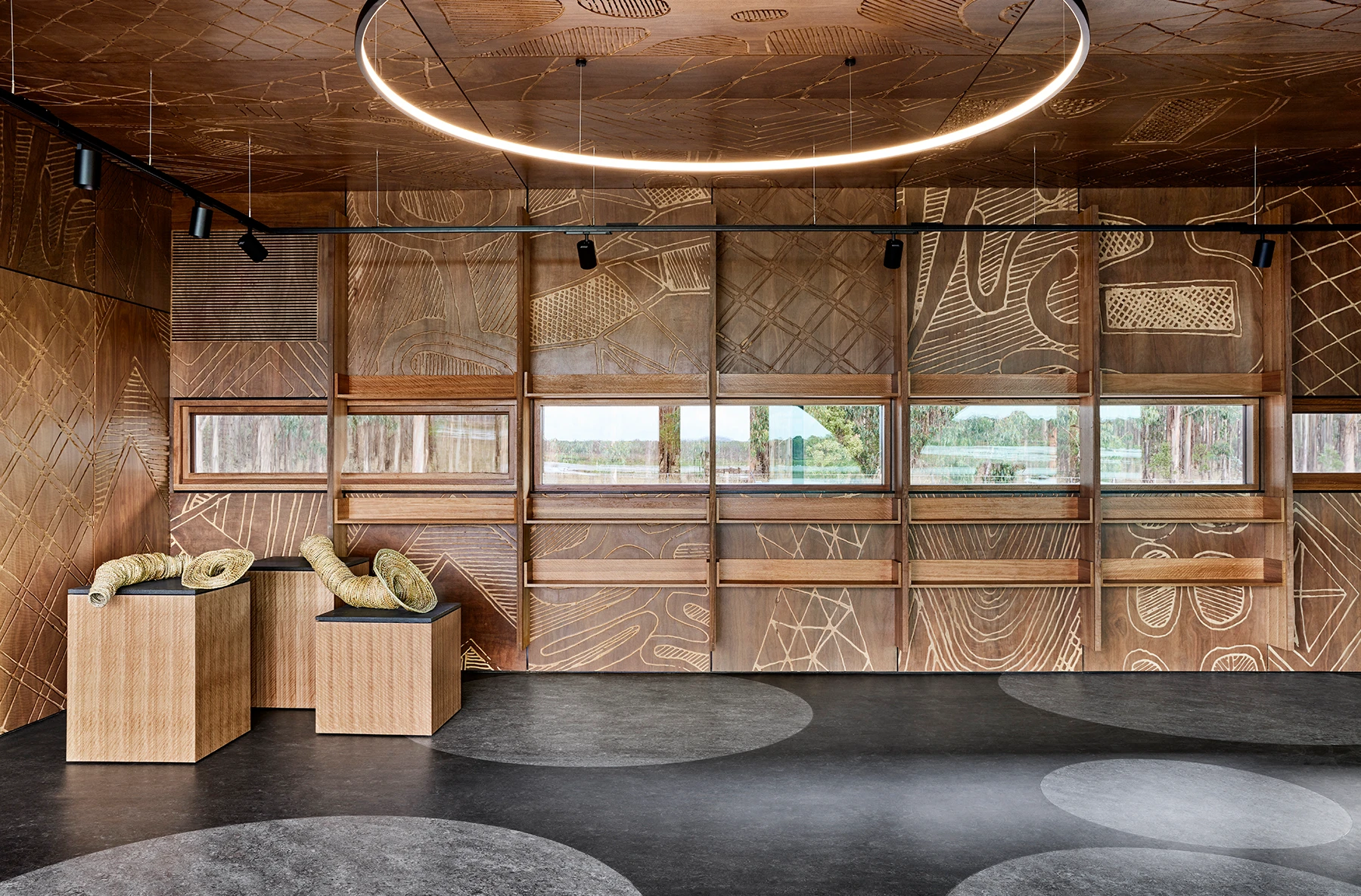 Modern interior with textured, patterned wood walls, a large circular ceiling light, and rectangular windows offering an outside view. Two woven benches and cylindrical cushions sit on the left atop a dark floor with circular patterns.