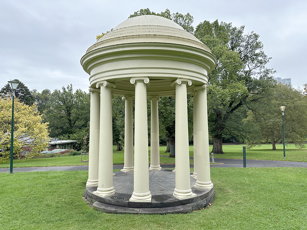 A small, round, cream-colored pavilion with a domed roof and eight columns stands on a grassy lawn in a park, surrounded by trees and paths under a cloudy sky.