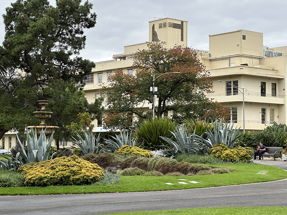 A landscaped garden with large agave plants, yellow shrubs, and a decorative fountain stands in front of a beige multi-story building. Two people sit on a bench; the sky is overcast.