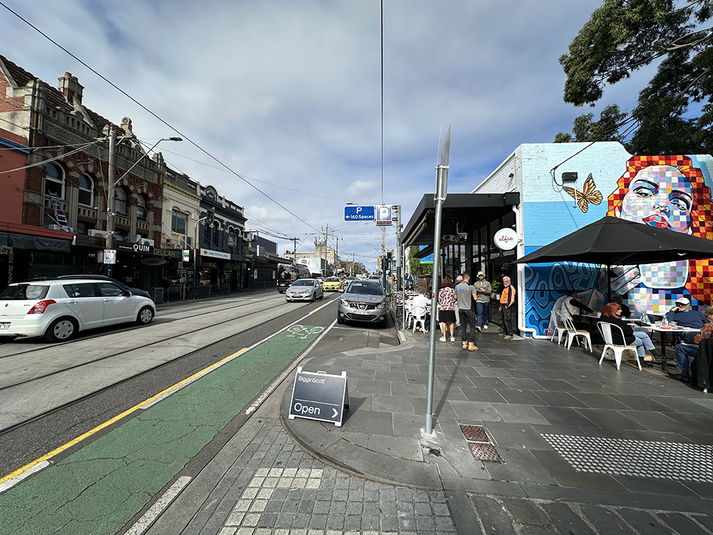 A busy urban street with shops, parked cars, and people sitting at outdoor tables. A colorful mural of a womans face decorates a building on the right. The sky is partly cloudy, and a bike lane runs alongside the street.
