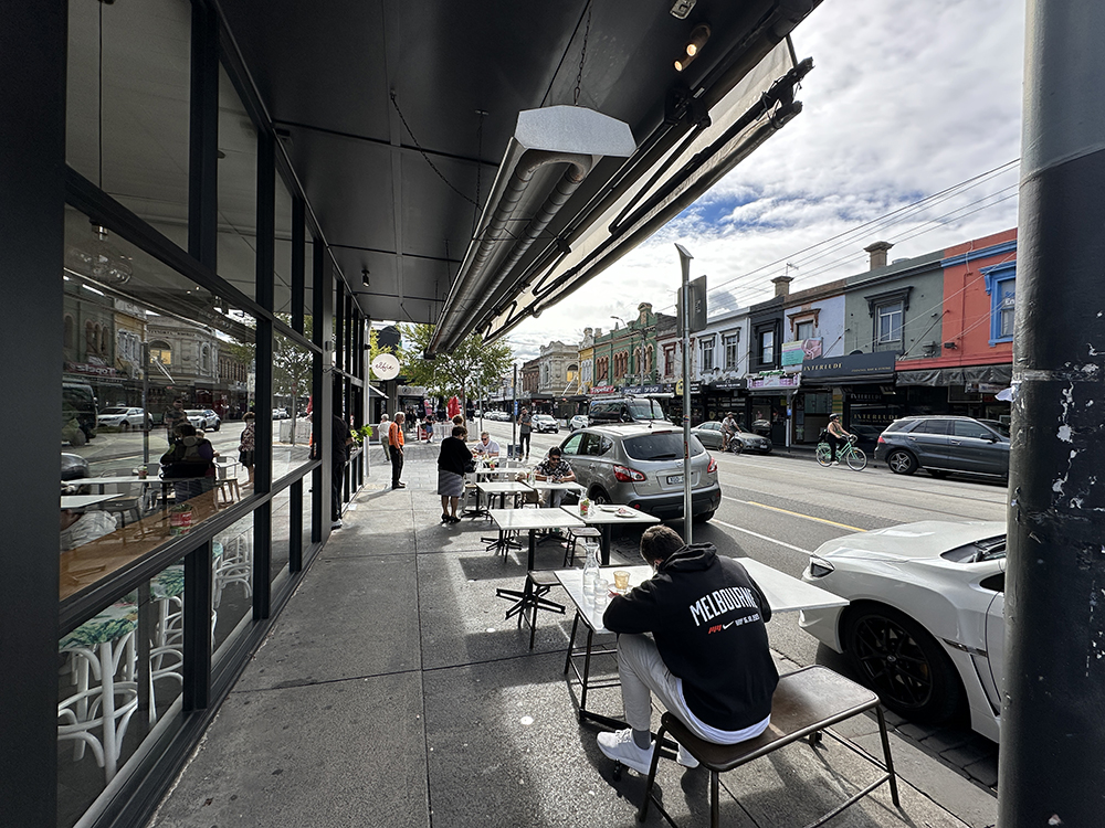People sit at outdoor tables along a city sidewalk next to a café. Cars are parked on the street, pedestrians walk by, and colorful shops line the opposite side under a partly cloudy sky.