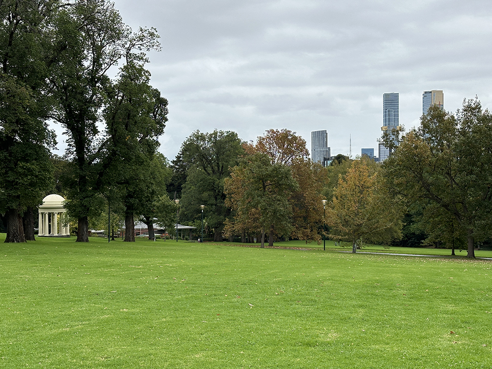A large green lawn bordered by trees with autumn foliage, a small domed pavilion on the left, and modern city skyscrapers visible in the background under a cloudy sky.