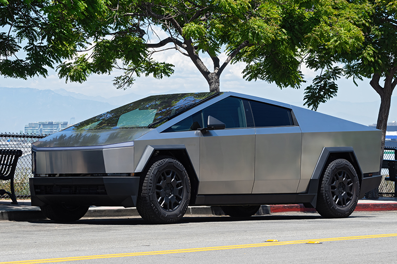 A silver Tesla Cybertruck is parked on a street near a sidewalk, with trees and a fence in the background on a sunny day.