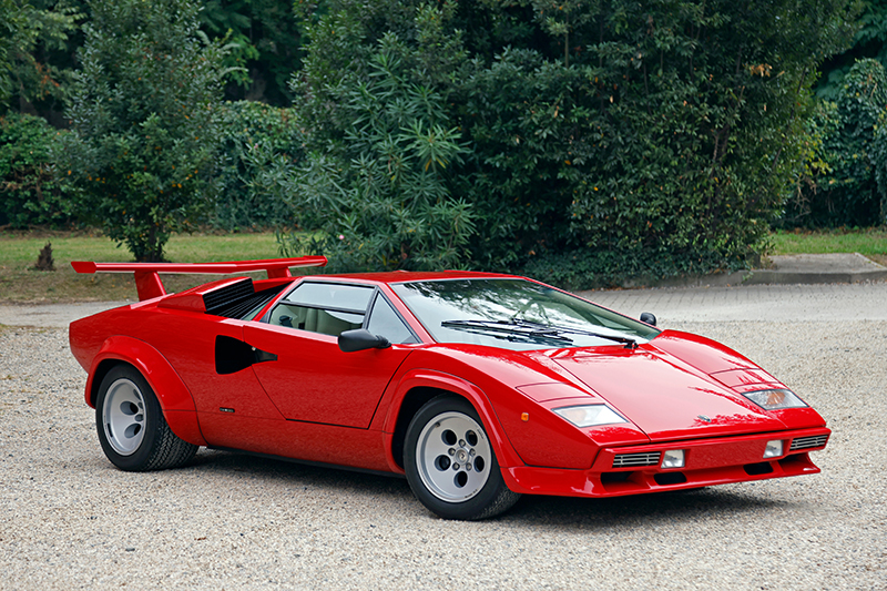 A bright red Lamborghini Countach sports car is parked on a gravel surface, surrounded by green bushes and trees in the background. The car has a low, angular design and a large rear wing.