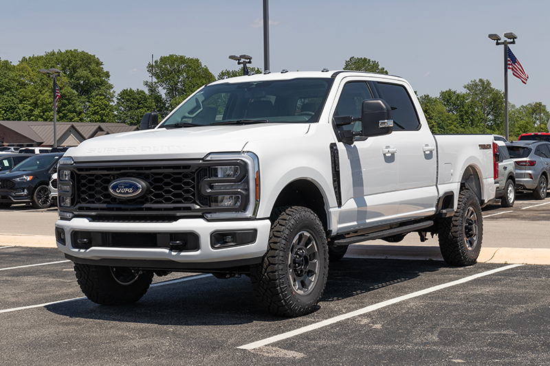 A white Ford Super Duty pickup truck is parked in a dealership lot on a sunny day, with trees and an American flag visible in the background.