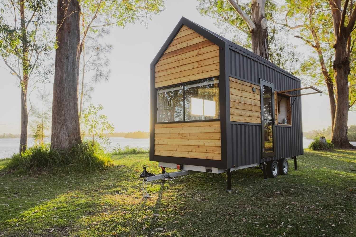 A modern tiny house on wheels sits on grassy ground near a lake, surrounded by tall trees. The house features wood and dark metal paneling, with large windows reflecting the sunlight.
