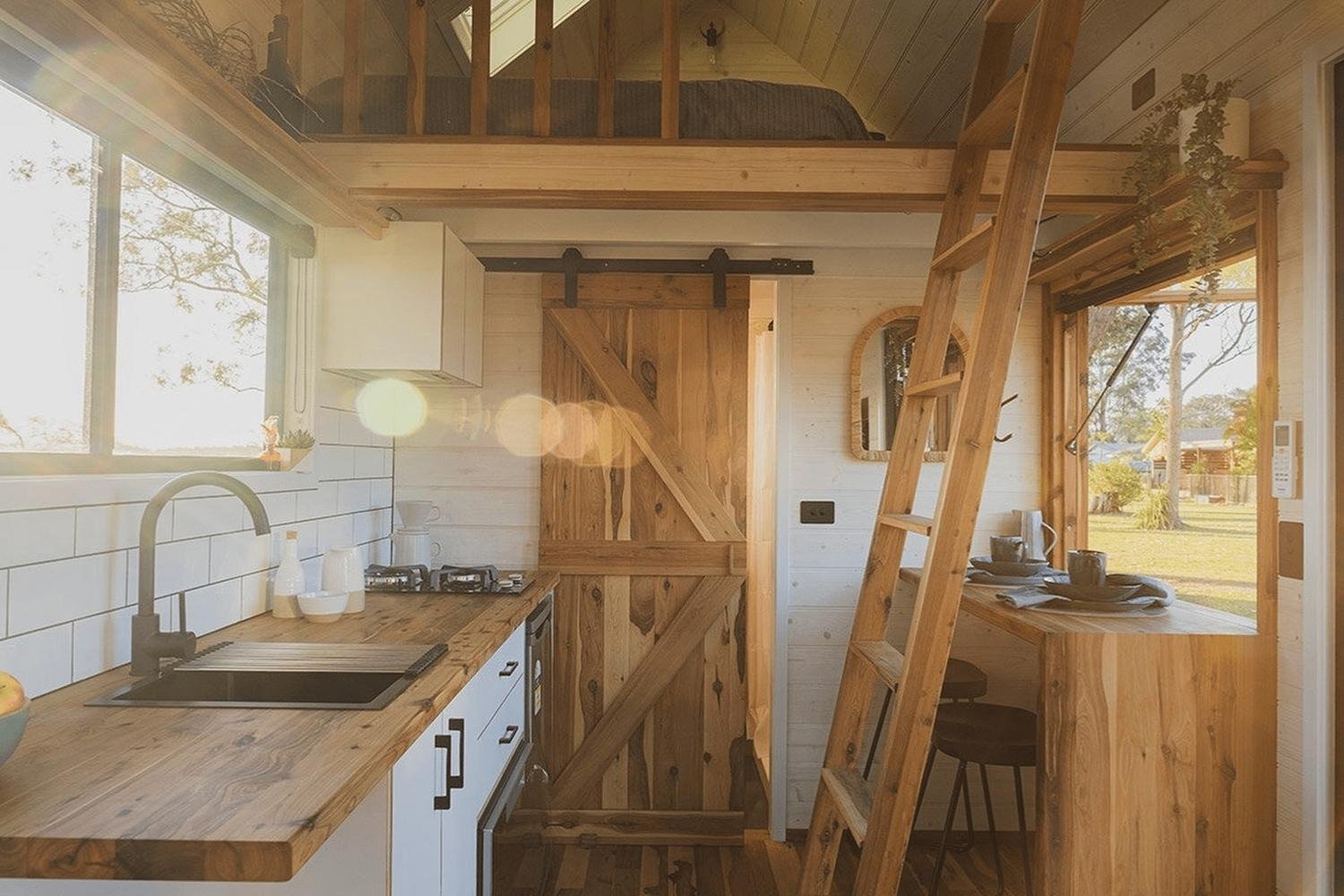 A cozy tiny house interior with wood accents, a small kitchen, a loft bed accessed by a ladder, a sliding barn door, and sunlight streaming through large windows.