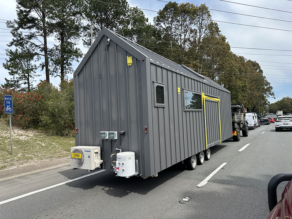 A gray tiny house on wheels is being towed along a road. The tiny house has several windows, solar panels on the roof, and an air conditioning unit attached at the rear. Cars and trees are visible in the background.