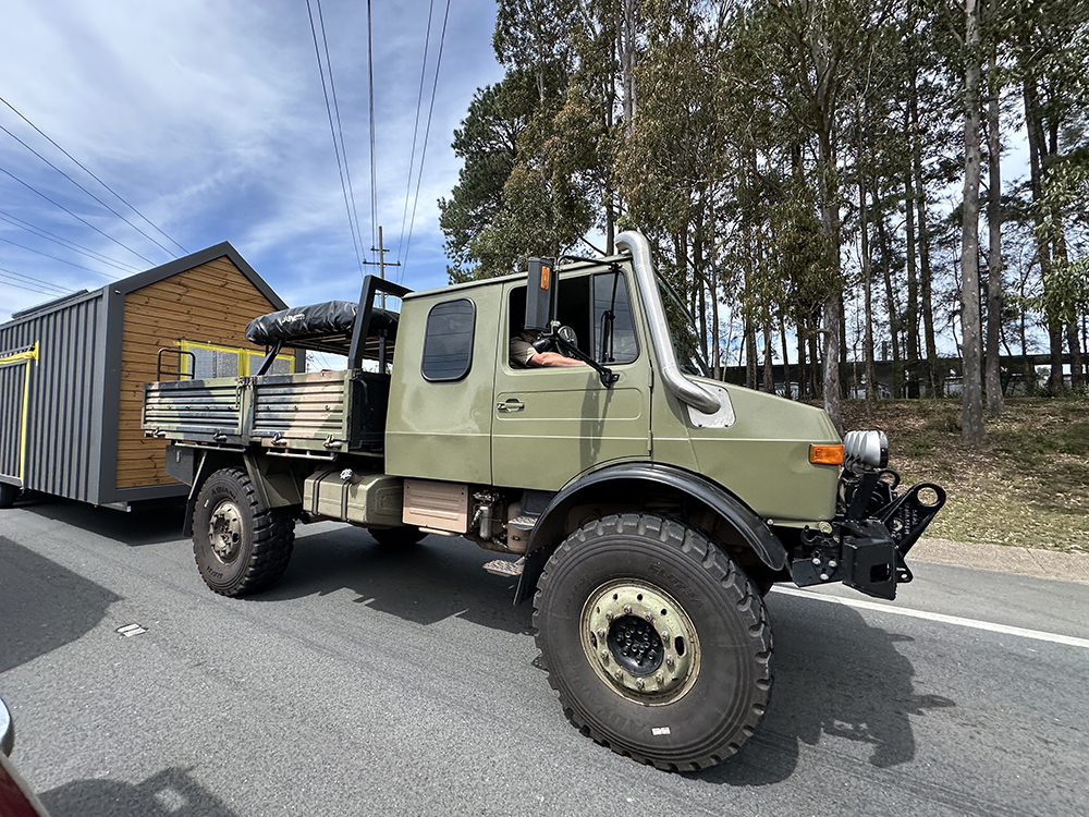 An olive-green off-road utility truck with large tires is driving on a road, pulling a trailer with a small house structure. There are trees and power lines in the background.