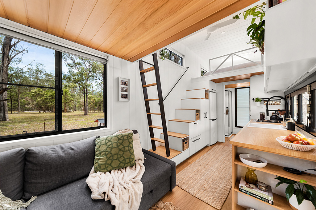 A modern tiny house interior with wooden floors and ceiling, a gray sofa, a cozy green pillow and throw, a loft with a ladder, large windows showing trees outside, and a compact kitchen with open shelves.