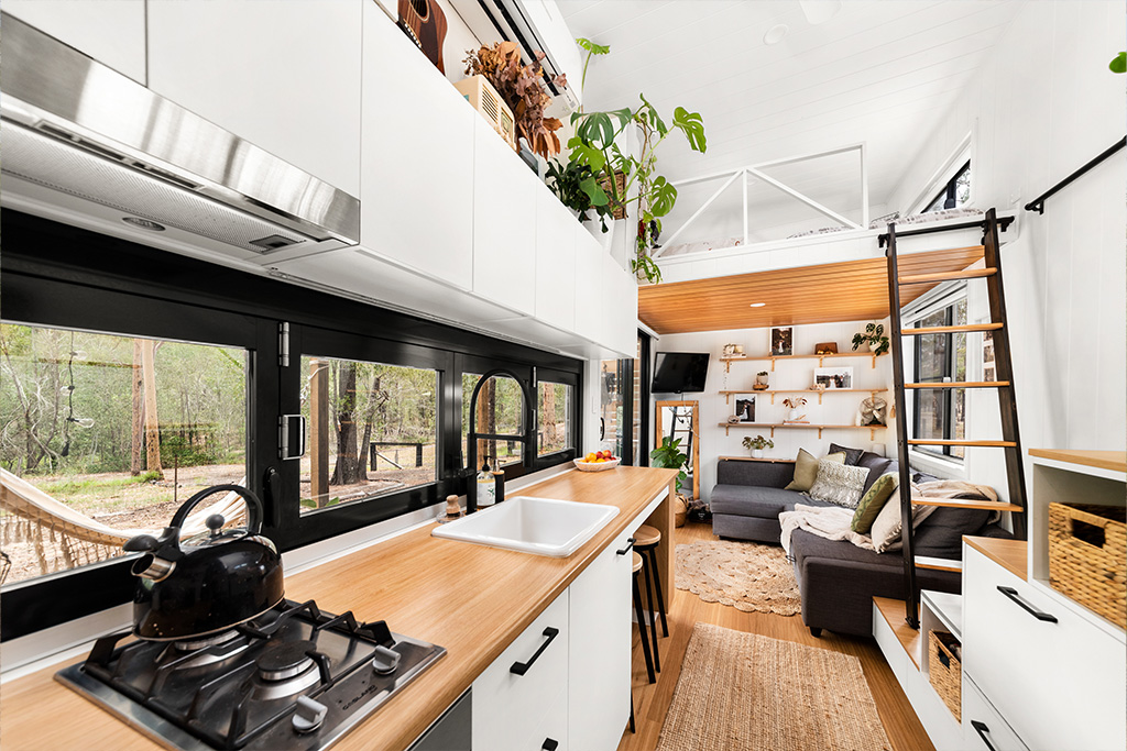 Modern, bright tiny house interior with a kitchen featuring white cabinets, wood countertops, and black fixtures. A cozy living area, ladder to loft, plants, and large windows with forest views complete the space.