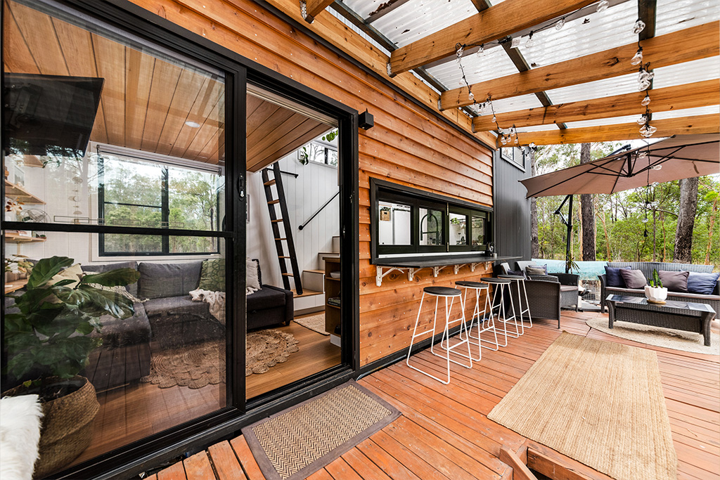 A cozy wooden patio with a bar counter and stools, large sliding glass doors, a covered pergola, and an outdoor seating area with a couch and umbrella beside a modern tiny house in a wooded area.