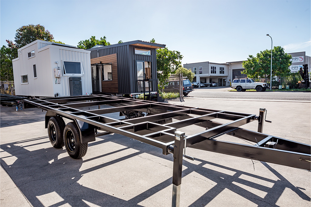 A black metal trailer frame on wheels is parked on pavement, with two completed tiny houses and modern buildings visible in the background under a clear sky.