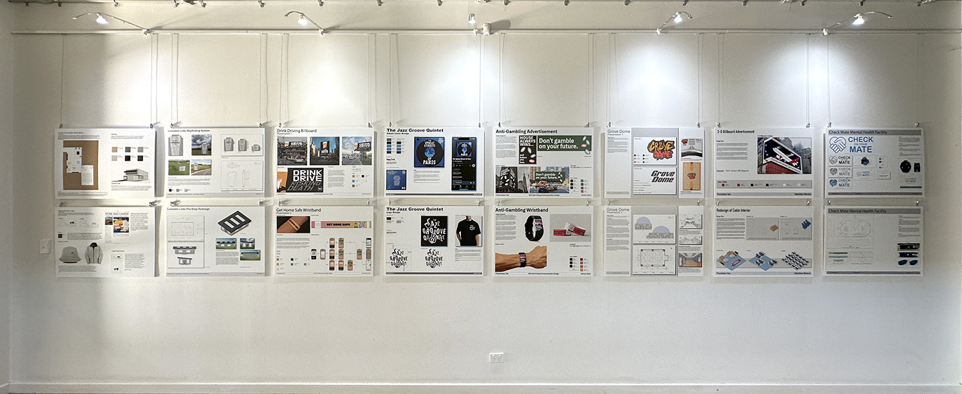 A white gallery wall displays 14 mounted posters in two rows, each showcasing design projects with images, text, and graphics under focused ceiling lights.