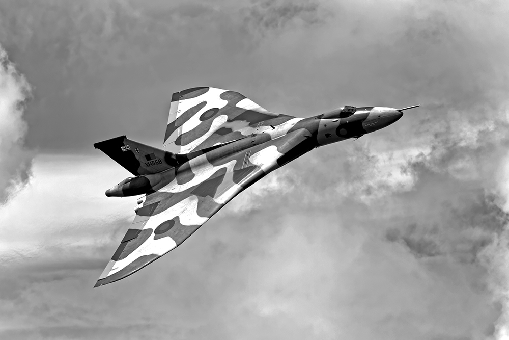 A camouflaged military jet, likely a bomber, flies at an angle against a cloudy sky. The aircraft has a distinctive delta wing design and a mottled paint pattern. The image is in black and white.