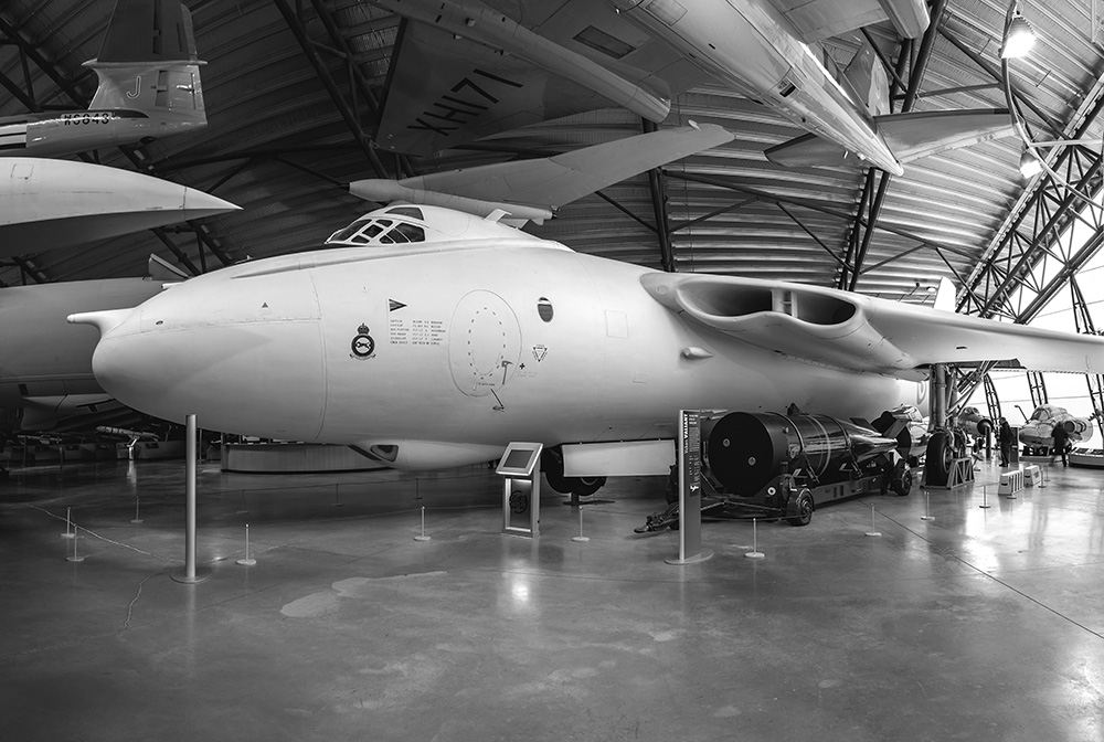 A large military aircraft is displayed inside a museum hangar, surrounded by information panels and barriers. Other aircraft and aviation exhibits are visible in the background. The photo is in black and white.