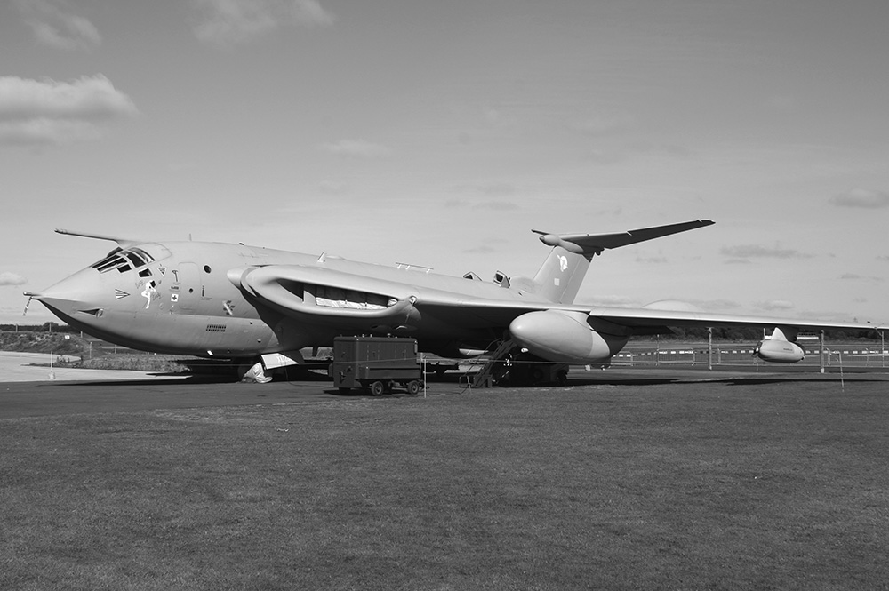 A large military aircraft with a distinctive angled nose and swept-back wings is parked on an airfield, with maintenance equipment nearby. The image is in black and white.