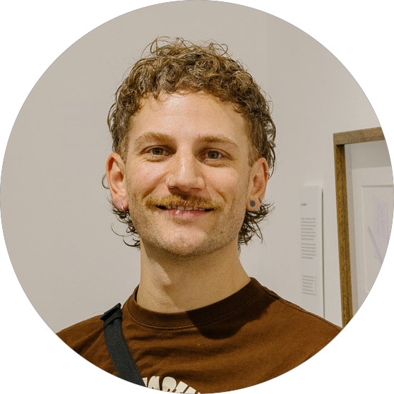 A person with short curly hair, a mustache, and earrings smiles at the camera. They are wearing a brown shirt and have a black strap over one shoulder. The background is indoors with framed art on the wall.