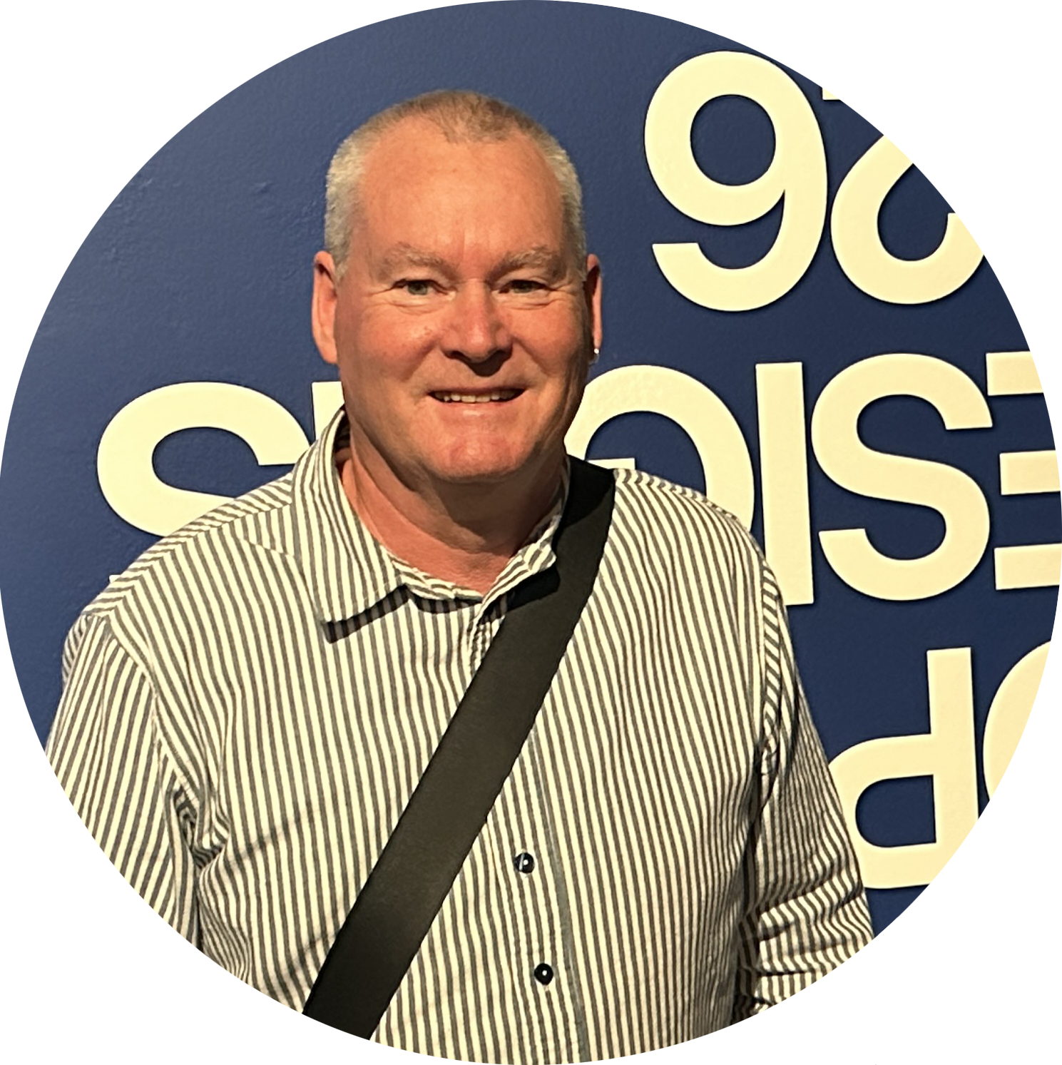 A man with short gray hair, wearing a striped button-up shirt and a black shoulder bag, smiles in front of a blue wall with large, partially visible white text.
